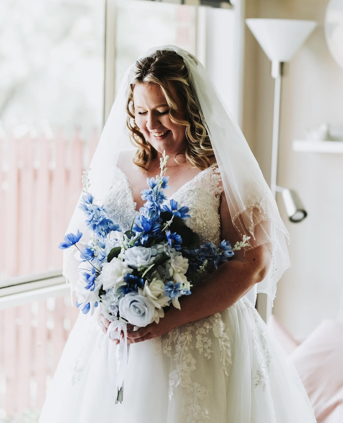 Bride holding a blue and white bridal bouquet featuring roses and daisies, wearing a white lace wedding dress and veil by a window in Canberra.