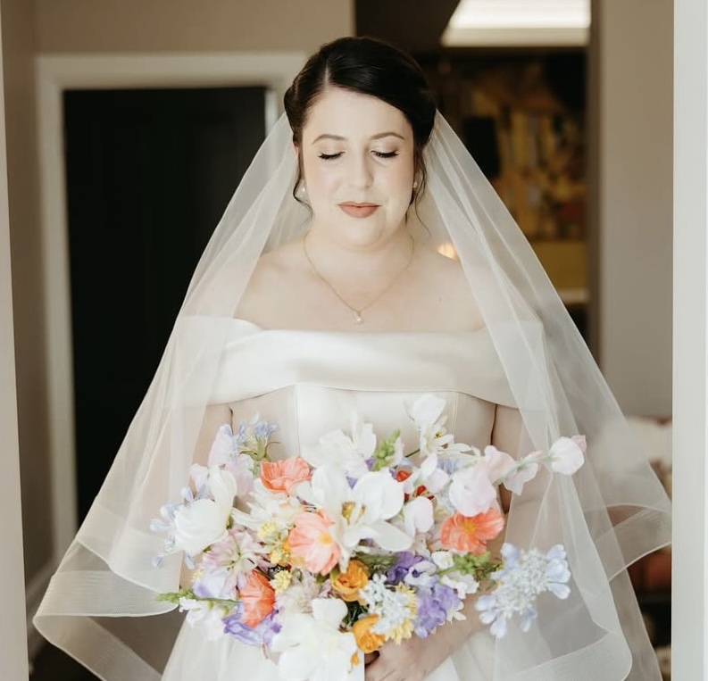 Bride in off-shoulder wedding dress holding colorful bridal bouquet with white, coral, and purple flowers, wearing veil and delicate necklace.