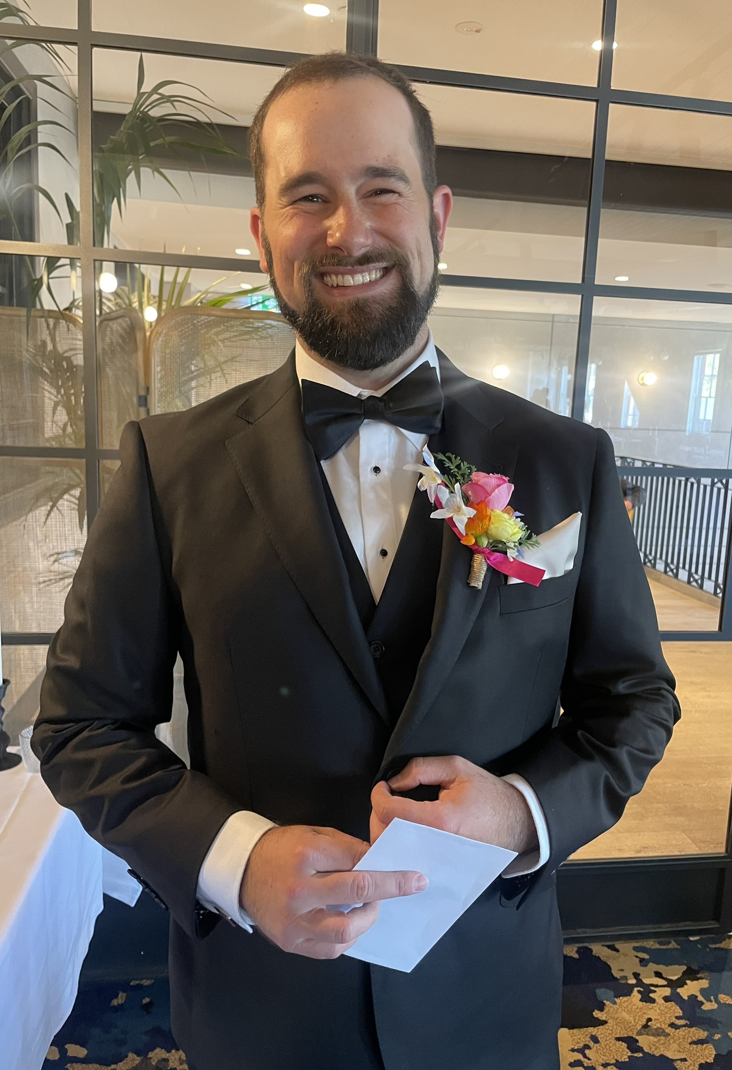 Groom in black tuxedo with colorful boutonniere smiling at Canberra wedding venue, holding ceremony program.